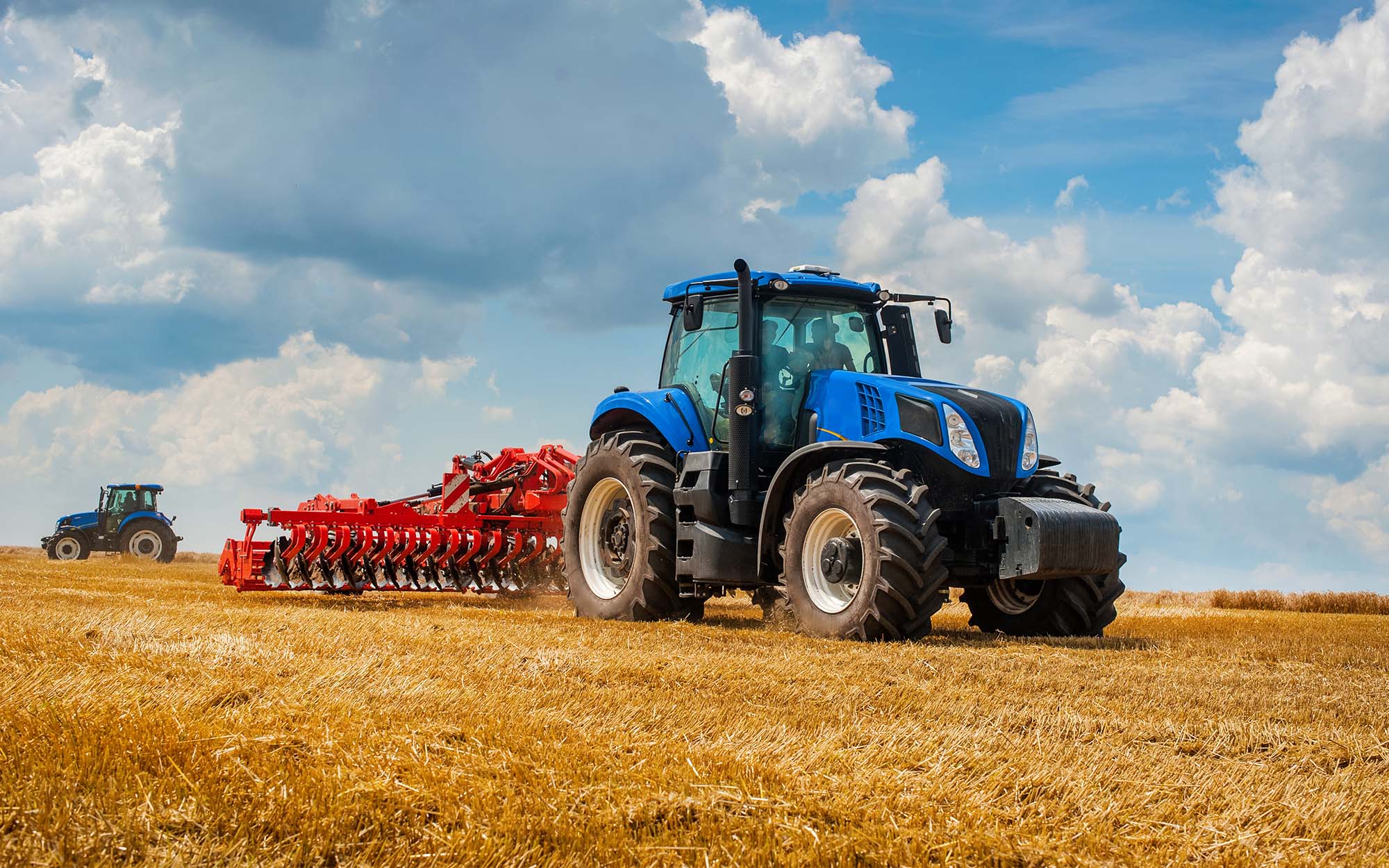 blue new tractor with red harrow in the field against a cloudy sky, agricultural machinery work